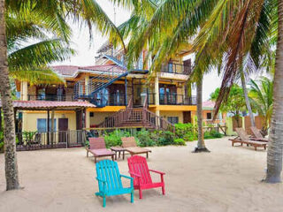 A beachfront villa with multiple levels, surrounded by palm trees. In the foreground, there are several wooden lounge chairs and two colorful Adirondack chairs on the sandy beach.