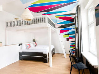 A bright, modern bedroom with a loft bed. Geometric, colorful patterns decorate the walls and ceiling. There are hardwood floors, a desk with a chair, and two windows with dark curtains.