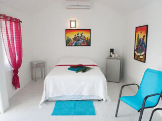 A small room with a bed covered in white linens, an artwork above the bed, a side table, a bright blue chair, red curtains, and a wall-mounted air conditioner.