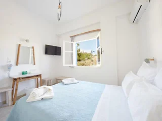 A small, bright bedroom with a neatly made bed, an open window, a wall-mounted TV, a vanity with a mirror, and an air conditioning unit. Two towels are folded on the bed.
