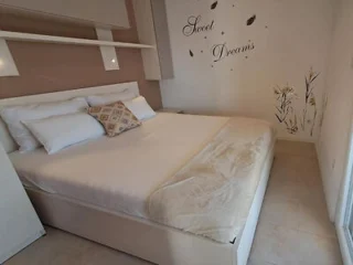A neatly made bed with white bedding and multiple pillows in a bright room. “Sweet Dreams” and decorative decals are on the wall above the bed.