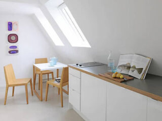 A bright kitchen with white cabinets, a small table with two chairs, framed wall art, and a skylight window. A cutting board with potatoes and an open book are on the countertop.