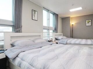 A bright bedroom with two neatly made beds, grey bedding, large windows with curtains, wall art, and a floor lamp in the corner.