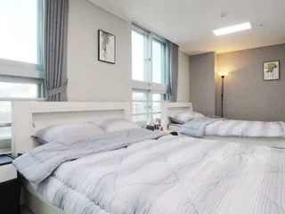A bright bedroom with two neatly made beds, grey bedding, large windows with curtains, wall art, and a floor lamp in the corner.