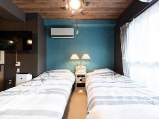 A cozy bedroom with twin beds, blue accent wall, and natural light streaming through the windows.