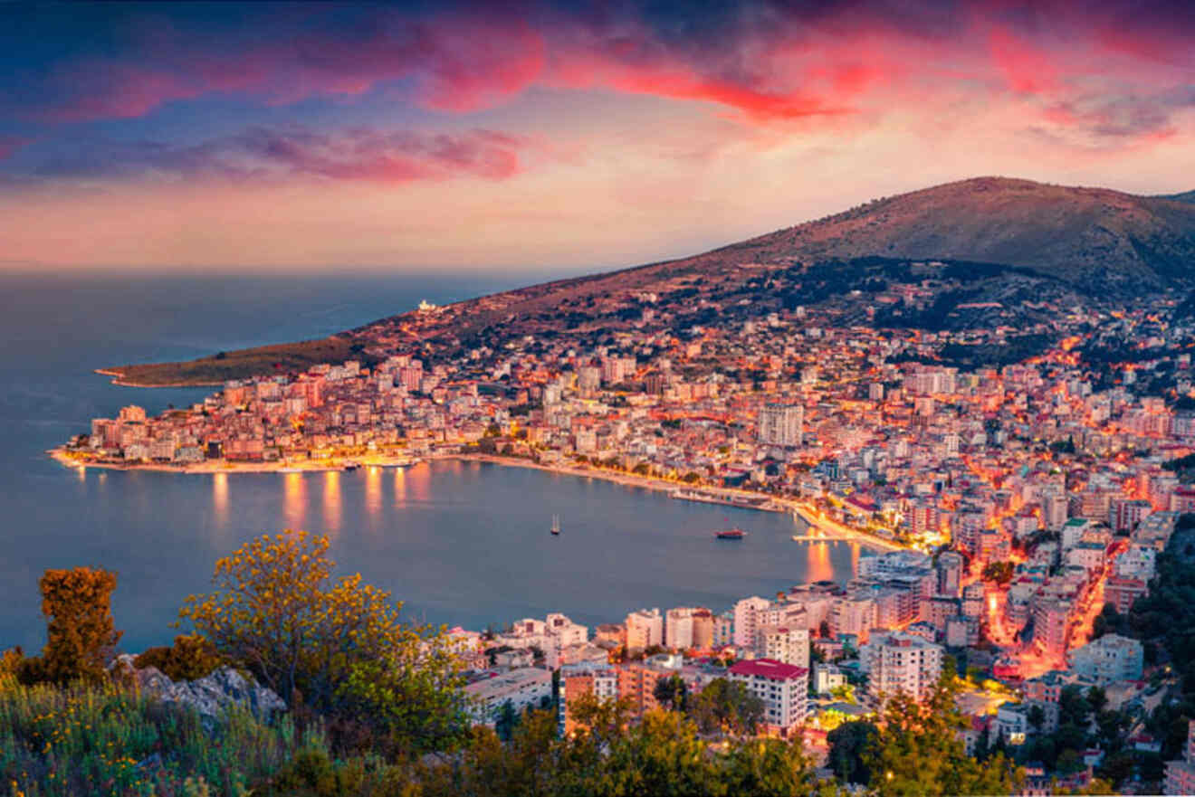 A coastal city at dusk, with illuminated buildings lining a bay, surrounded by hills and under a vibrant pink and purple sky.