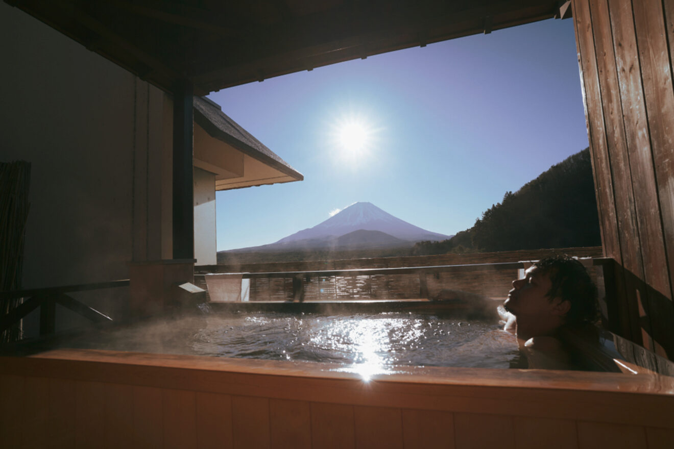 A person relaxing in a hot spring bath with a stunning view of Mount Fuji and the sun shining above.