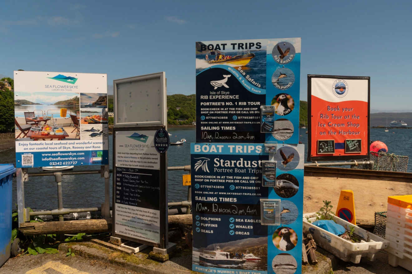 Information boards and signs by the waterfront advertising boat trips and tours. Small boats and buoys are visible in the background on a sunny day.