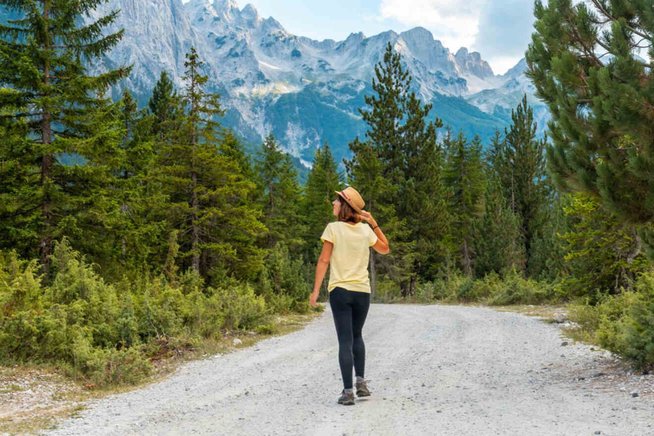 A woman in hiking gear walks along a dirt path surrounded by dense forest and towering mountains.