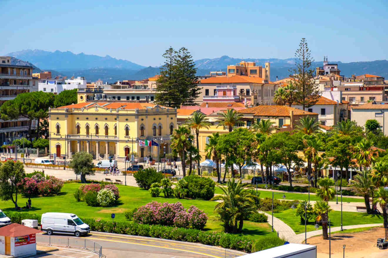 A sunlit Mediterranean town with historic buildings, green lawns, palm trees, parked vehicles, and mountains in the background.