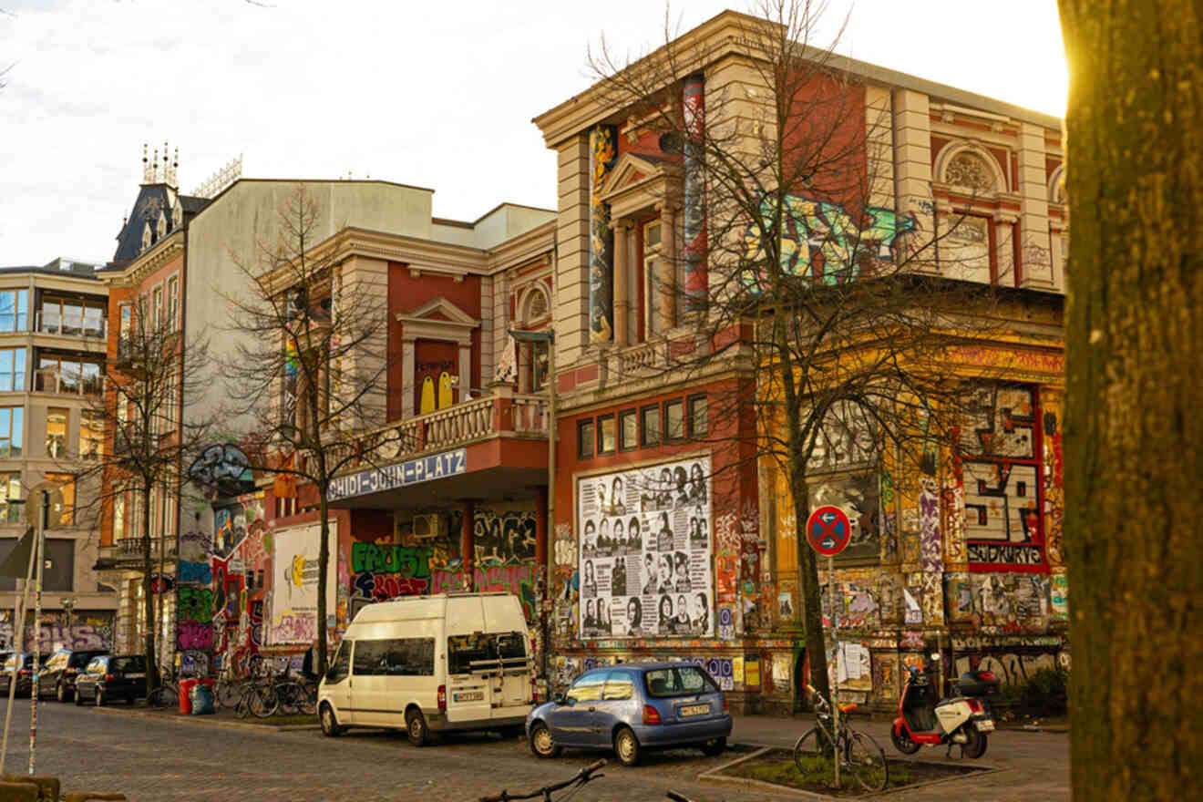 A street scene shows colorful graffiti and posters covering the walls of historic buildings. Several parked vehicles and bicycles are visible. Trees without leaves stand nearby.