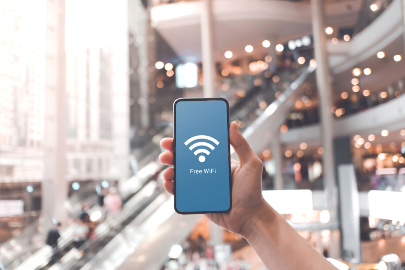 A hand holding a smartphone with a "Free WiFi" screen, set against a blurred background of a bustling indoor mall.