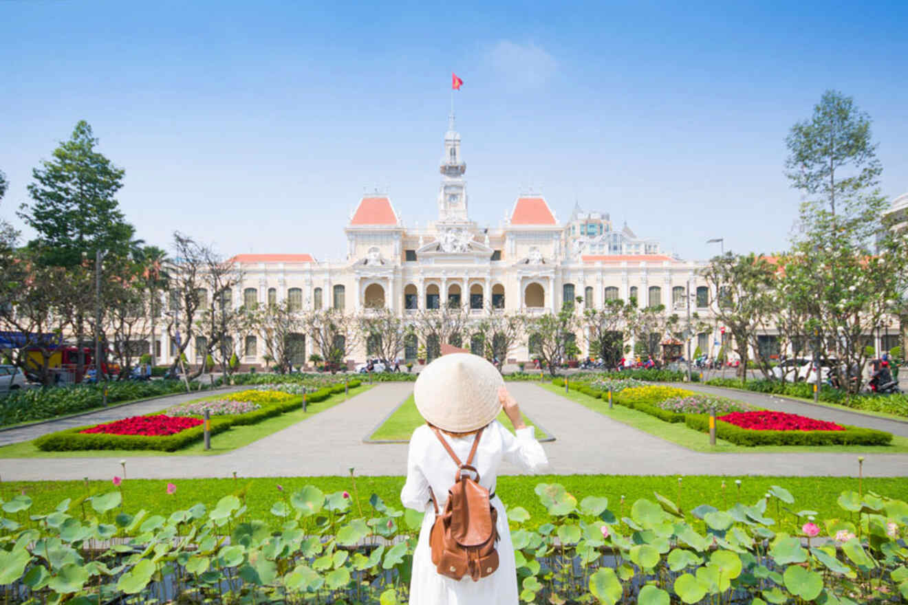 A person wearing a conical hat stands in a garden facing a grand, white colonial-style building with a red roof under a clear blue sky.