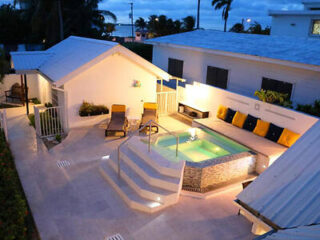 An aerial view of a modern backyard featuring a lit pool with adjacent lounge chairs, surrounded by white buildings and palm trees, all under an evening sky.