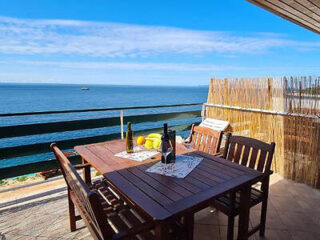 A wooden table with chairs and wine bottles on a balcony overlooks the ocean under a clear blue sky.