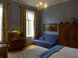 A bedroom with blue walls, a bed, a blue sofa, a wooden wardrobe, a desk, floral curtains, a chandelier, and a framed painting on the wall.