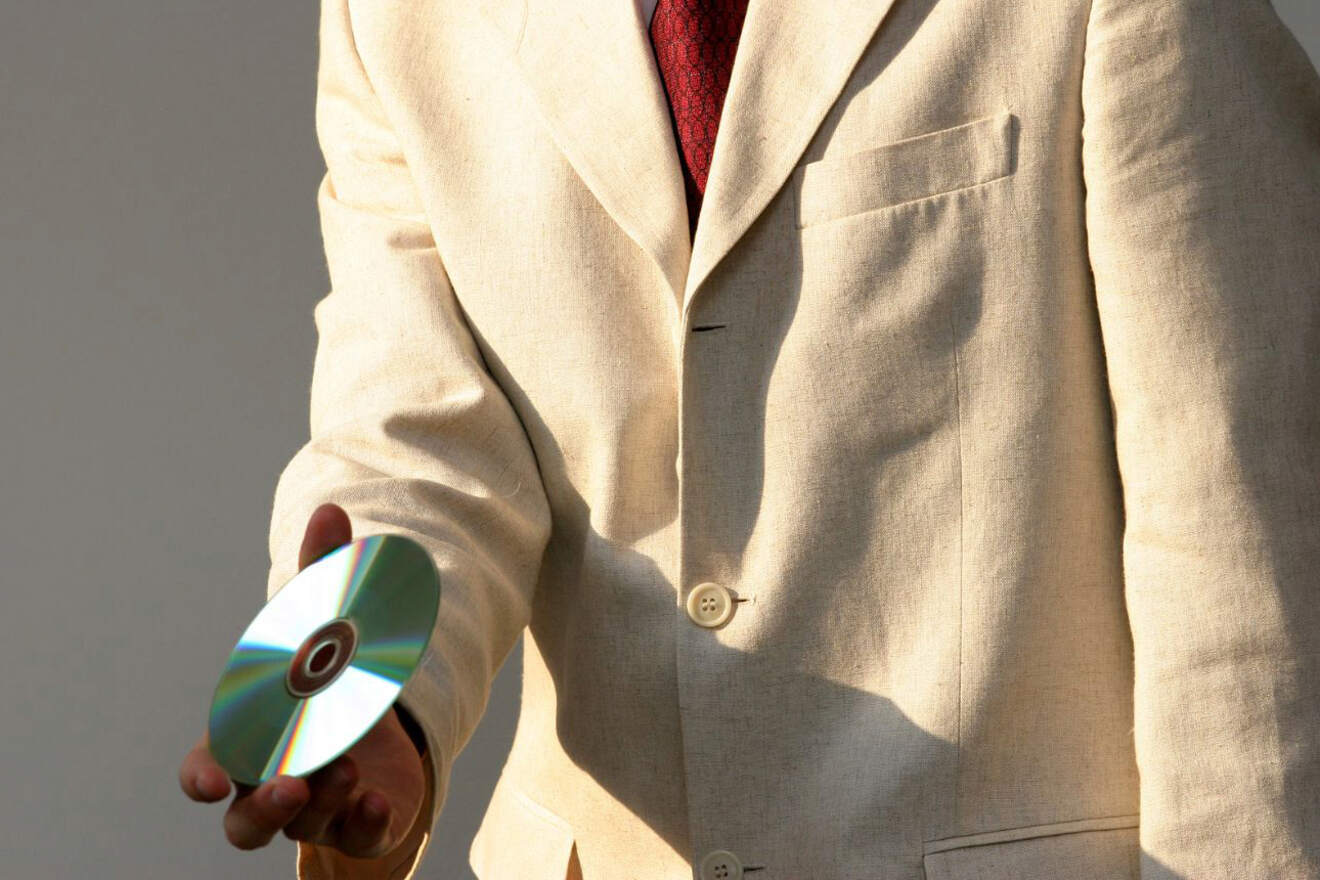 A person in a beige suit and red tie holds a CD in their hand, the CD reflecting light against a plain background.