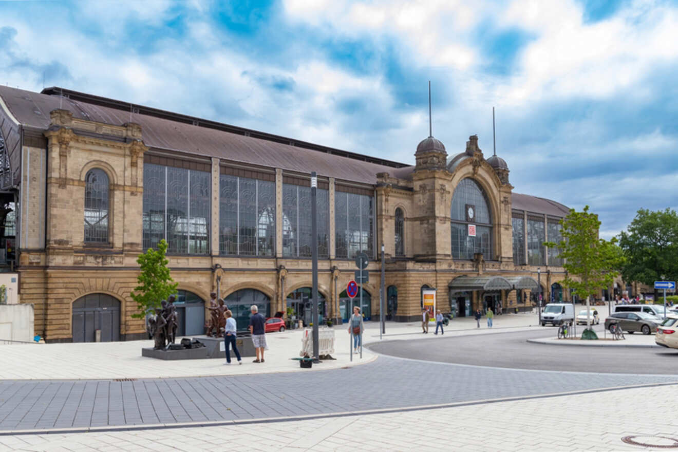 A historic train station with a large clock and arched windows, surrounded by people walking and standing, cars parked nearby, and a partly cloudy sky above.