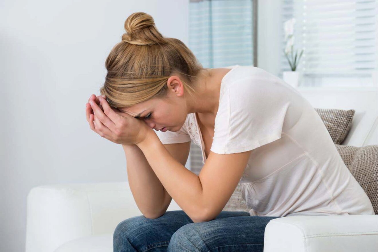 A woman sits on a couch with her head in her hands, appearing stressed.