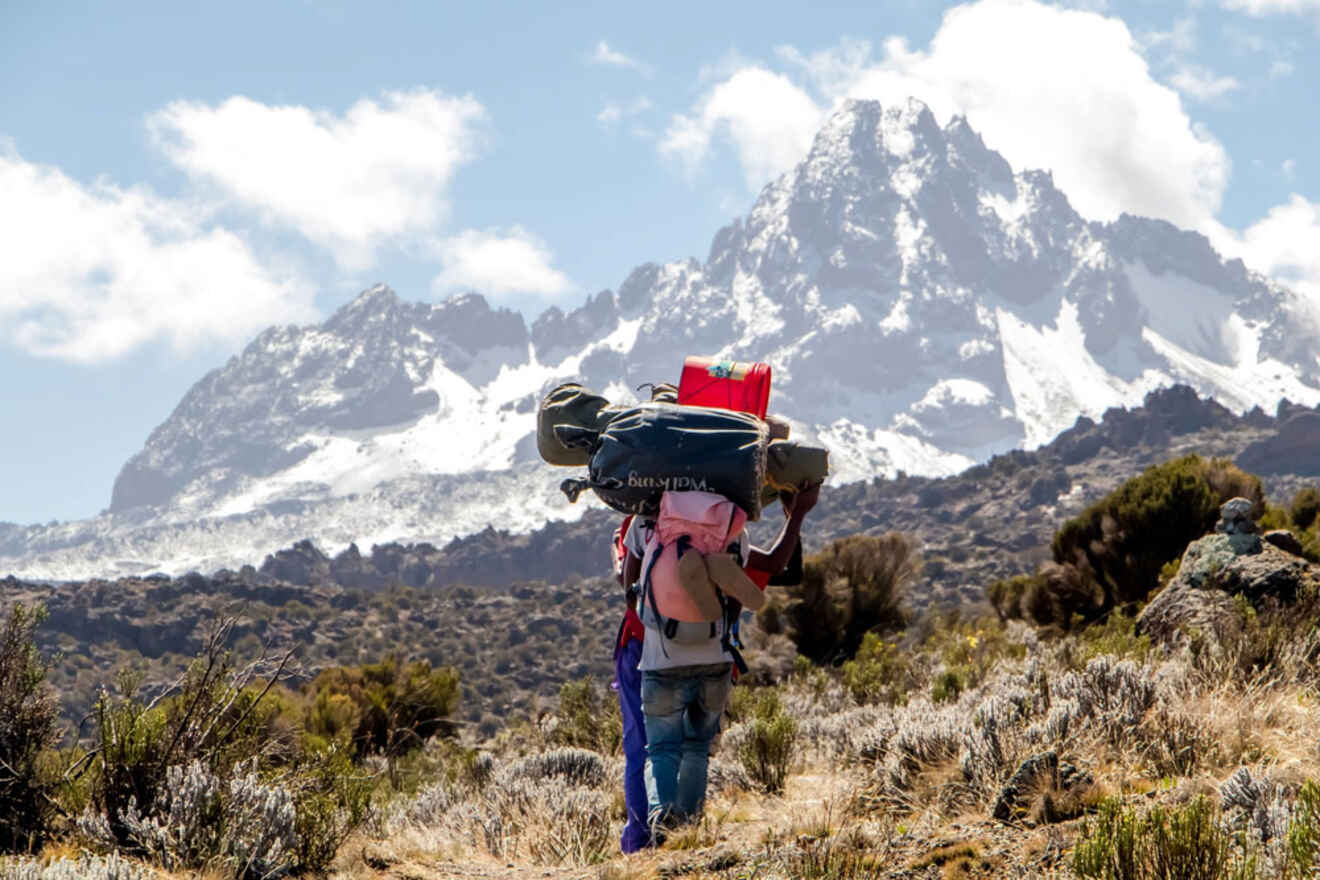 Two hikers with large backpacks walk on a trail toward a snowy mountain peak under a partly cloudy sky.