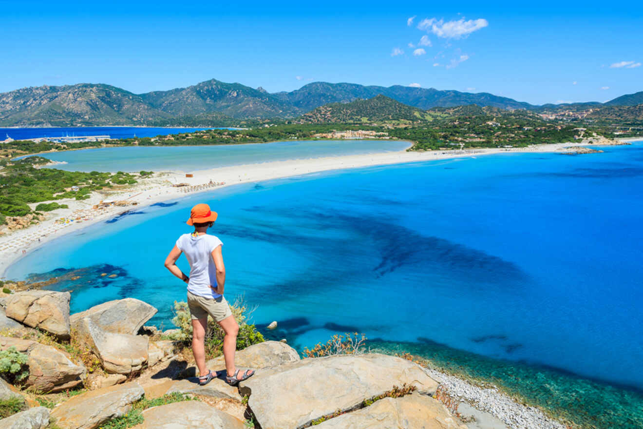 A person in a hat stands on a rocky outcrop overlooking a scenic beach with clear blue waters, green hills, and a sunny sky.