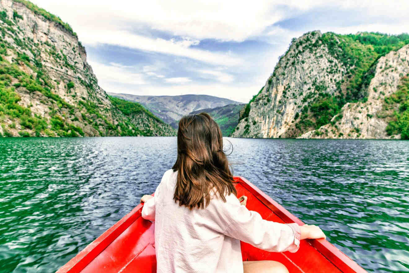 A woman in a red boat looks out at the serene waters of Komani Lake, framed by steep, green cliffs.