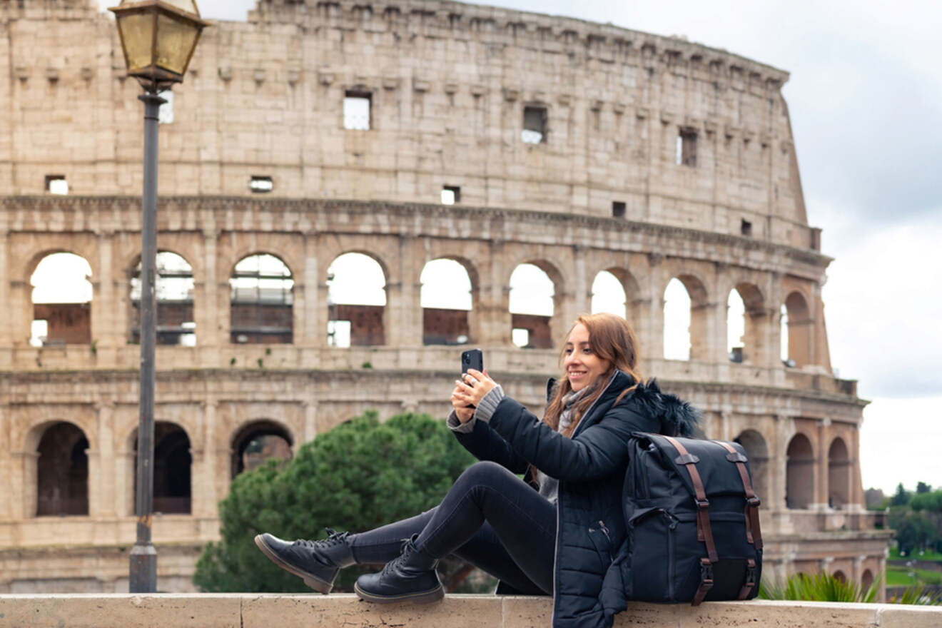 A person wearing a black jacket and carrying a backpack sits in front of the Colosseum, holding a smartphone. The ancient Roman amphitheater is prominent in the background.