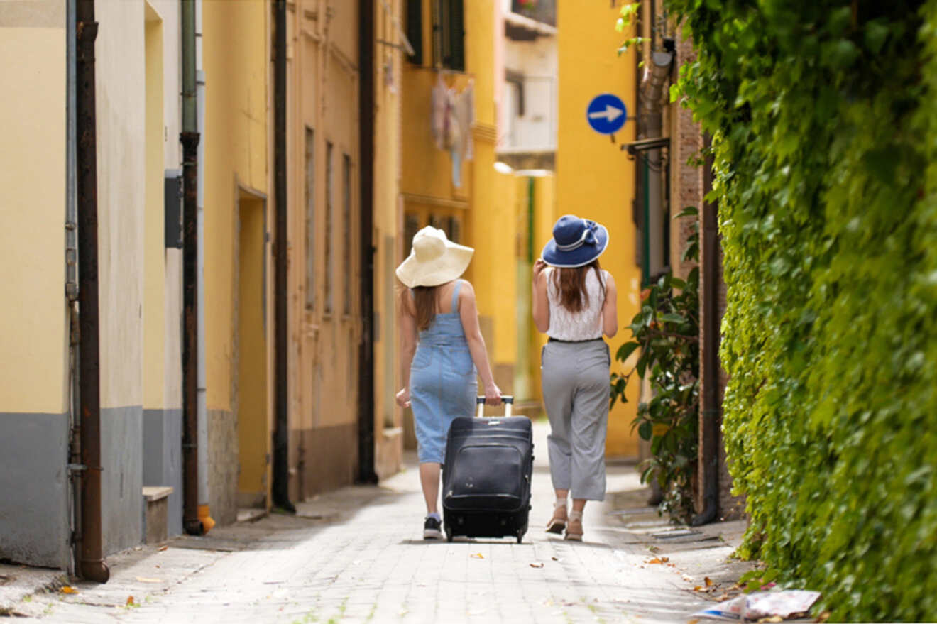 Two people in sun hats walk down a narrow, sunlit alley with luggage, flanked by yellow buildings and green foliage.