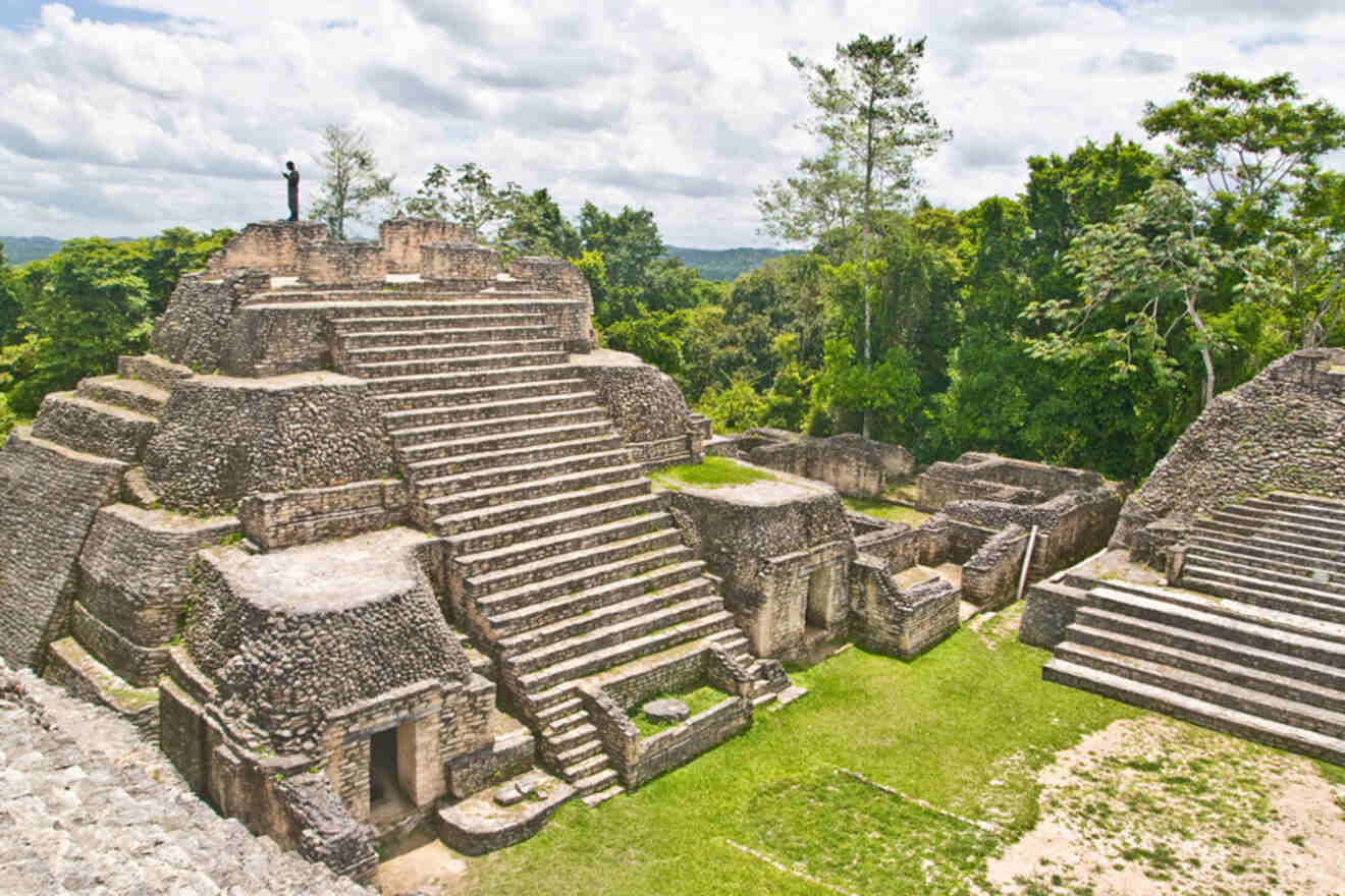 Ancient stone ruins of a stepped pyramid with surrounding structures, located in a lush green forest under a partly cloudy sky.