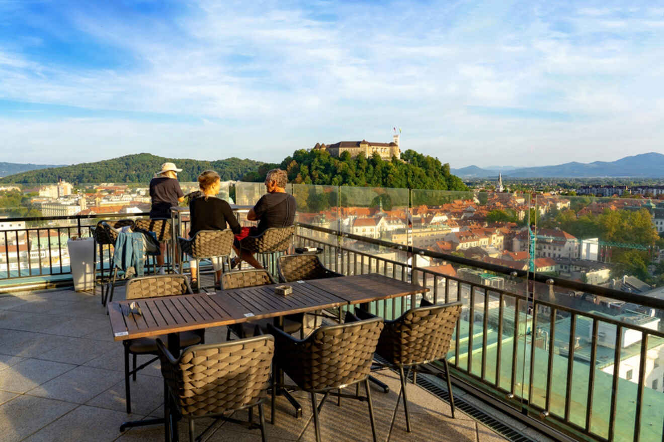 Three people stand around a table on a rooftop terrace, overlooking a cityscape with a hilltop castle in the distance under a clear sky. Empty chairs surround another table in the foreground.