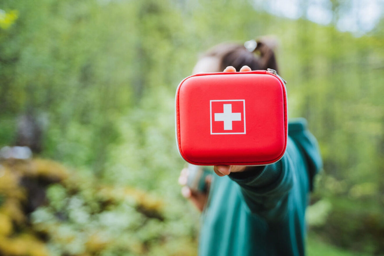 Person holding a red first aid kit in an outdoor setting with green foliage in the background.