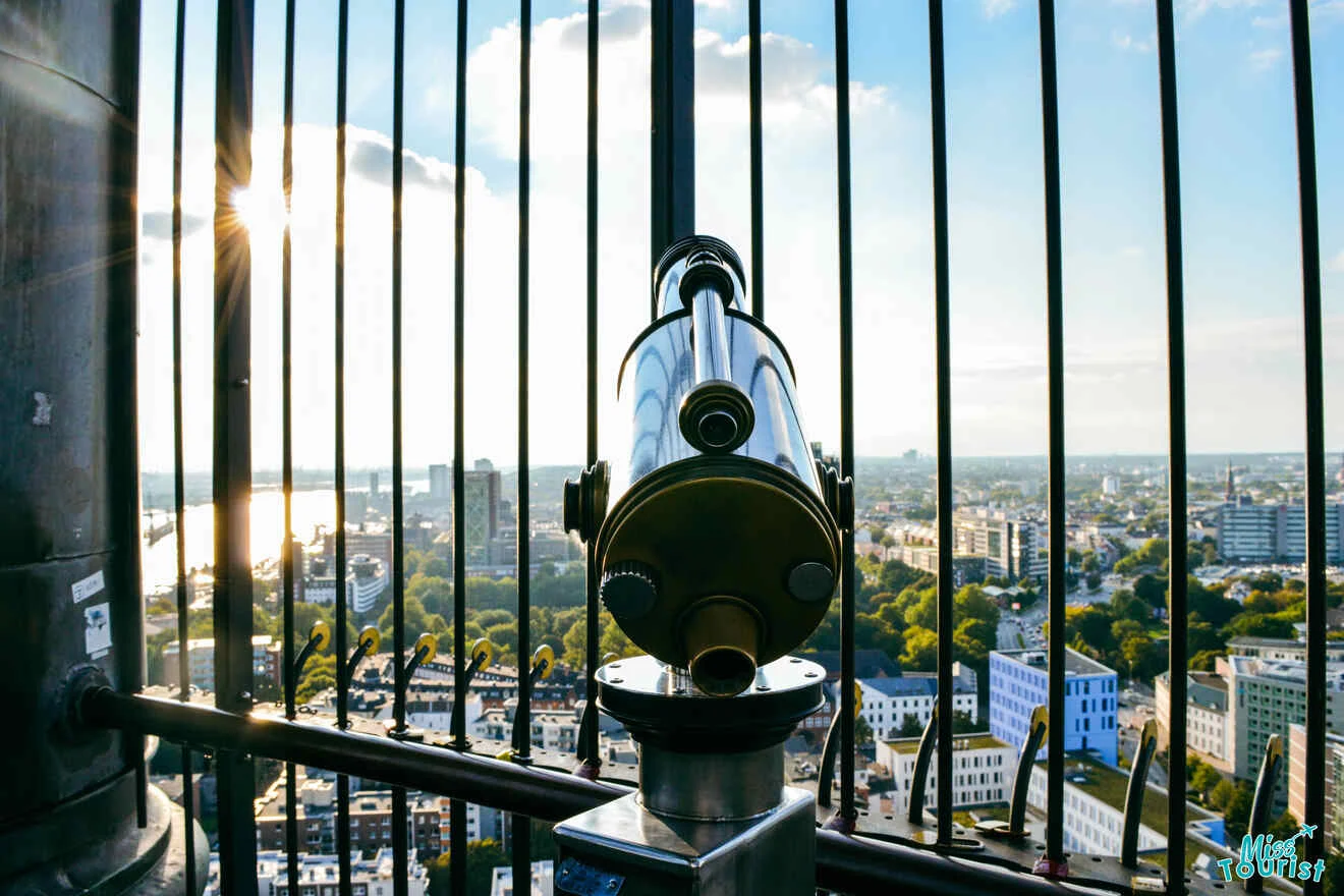 A metal telescope is mounted on a railing, facing a cityscape with buildings, greenery, and a river under a partly cloudy sky.