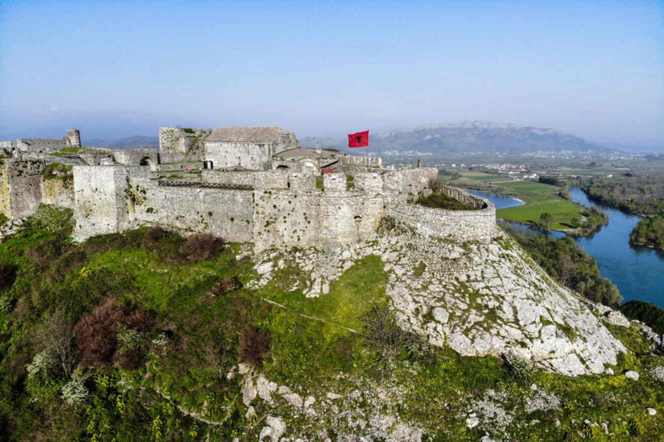 Aerial view of the historic Rozafa Castle in Shkoder, perched on a hill with a red flag flying and a river winding through the landscape.