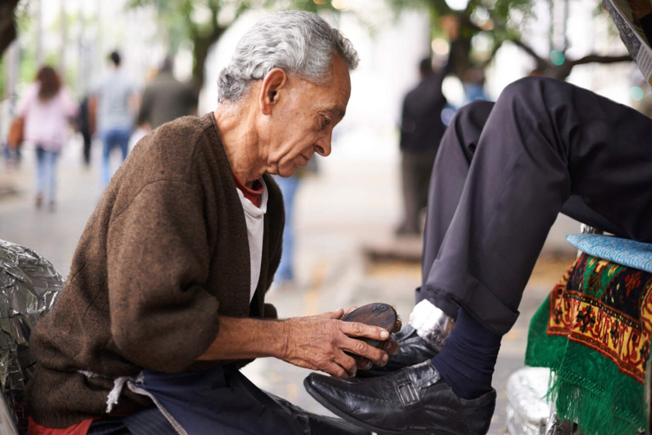 An elderly shoeshiner polishing a customer's shoe on the street, with a focused expression, surrounded by greenery and pedestrians in the background.