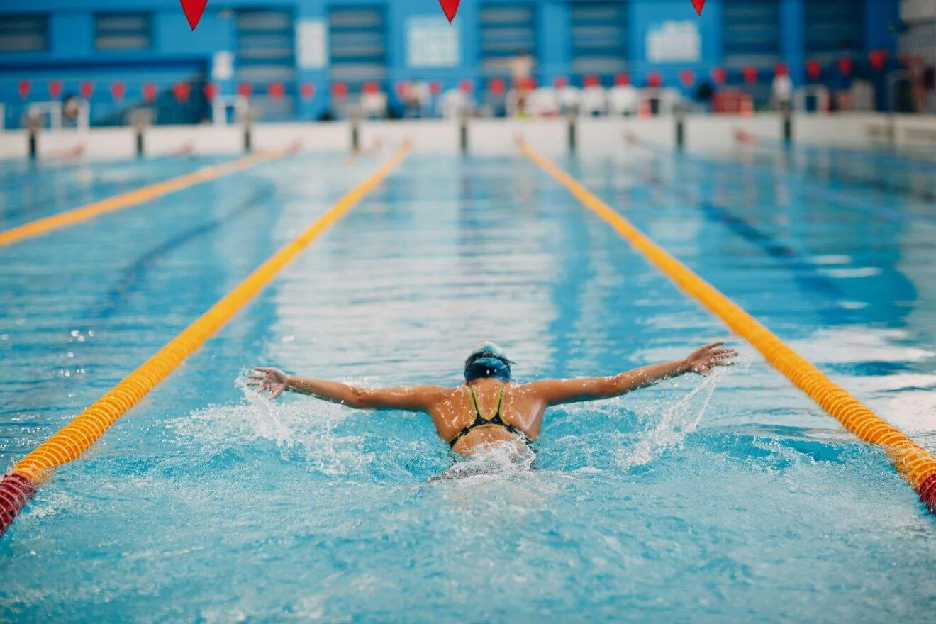 A swimmer performs a butterfly stroke in an indoor competition pool, with lane dividers on both sides and red flags overhead.