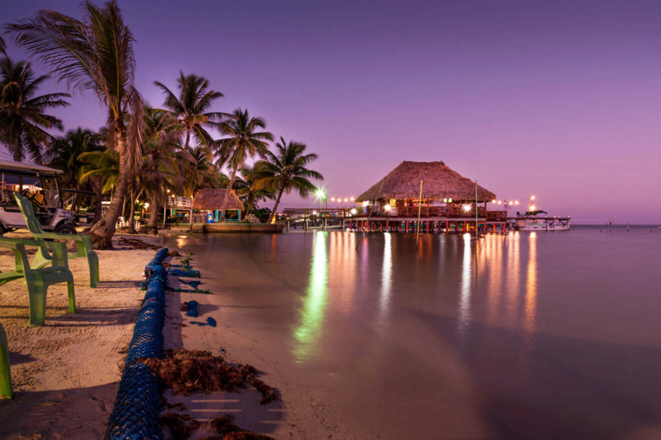 A beach at twilight with palm trees and a thatched-roof building over the water, illuminated by lights, reflecting on the calm sea. Nearby, plastic chairs and seaweed are visible on the sandy shore.