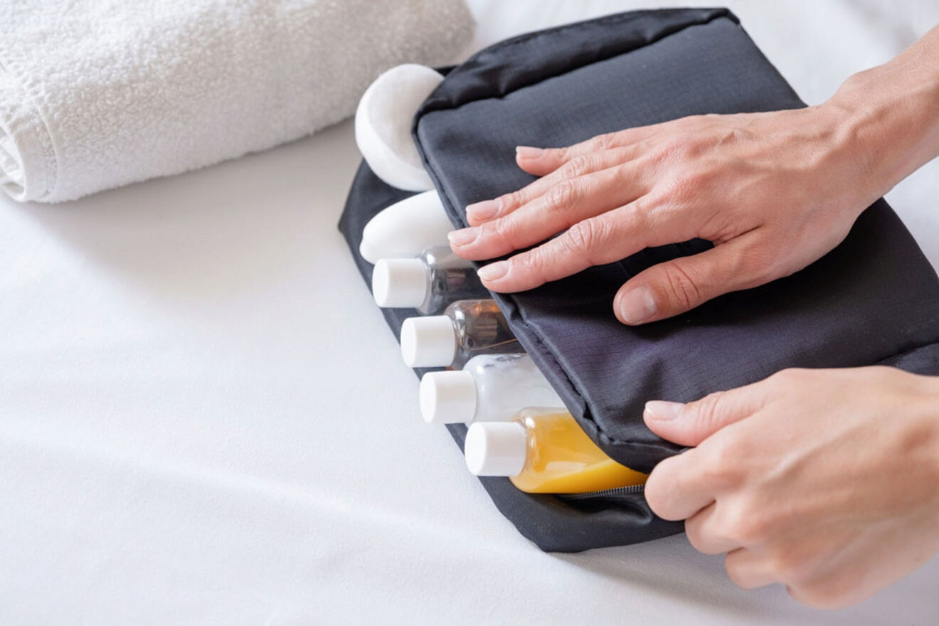 Person packing small bottles of toiletries into a black travel bag on a white surface, with a white towel and soap beside the bag.