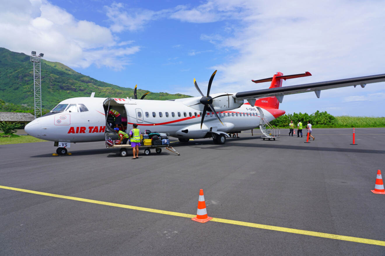 Air Tahiti plane being loaded with luggage on a sunny day, with lush green hills in the background.