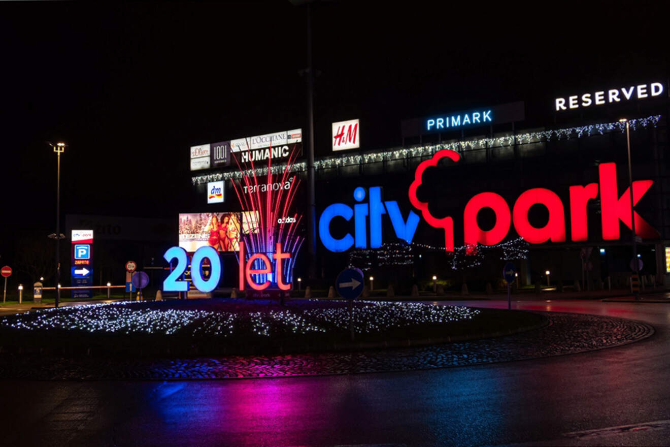 Night view of a shopping mall entrance decorated with bright, colorful signs including "city park" and a “20et” display with store logos like H&M, Primark, and Reserved visible in the background.