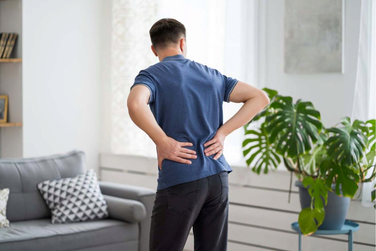 A man holding his lower back in pain while standing in a living room.