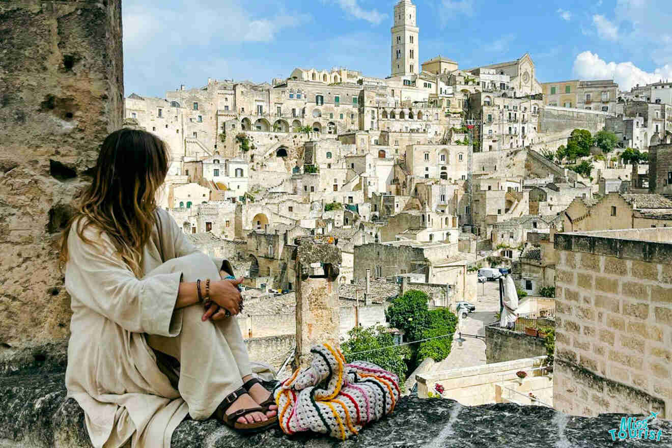 A person in a long-sleeved outfit sits on a rock wall overlooking a historic town with numerous stone buildings and a tall bell tower in the background.