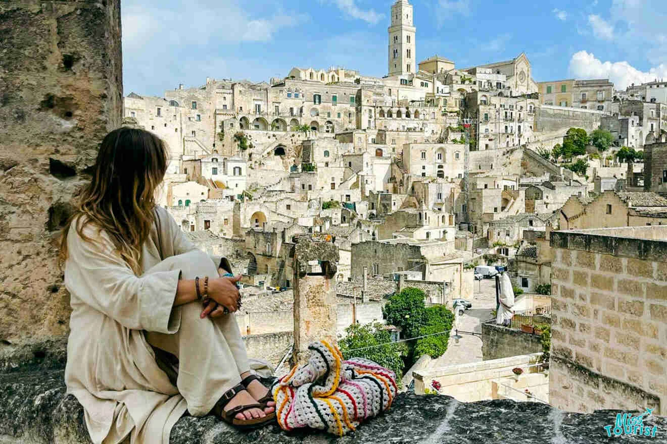 A person in a long-sleeved outfit sits on a rock wall overlooking a historic town with numerous stone buildings and a tall bell tower in the background.