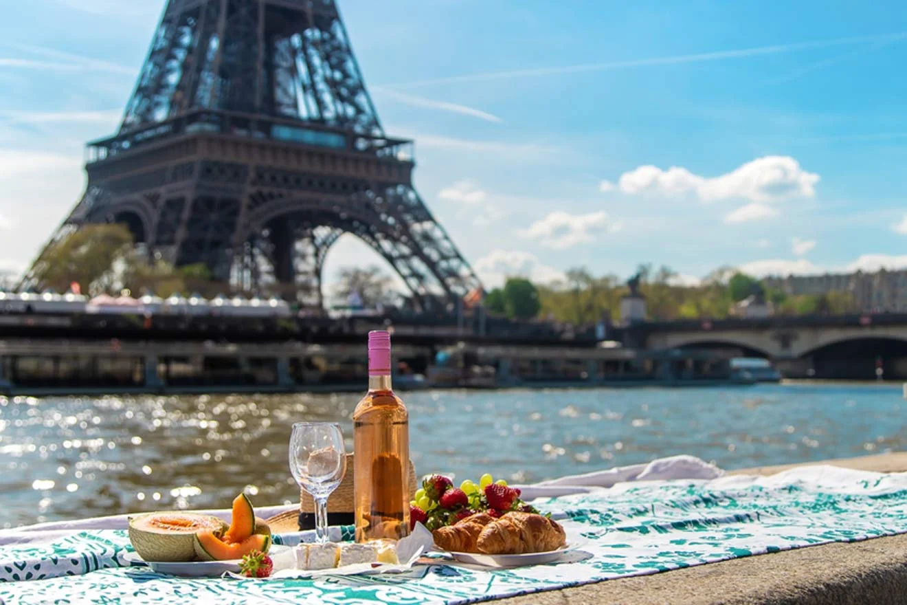 A picnic setup with wine, cheese, fruits, and a croissant on a riverside near the Eiffel Tower on a sunny day.