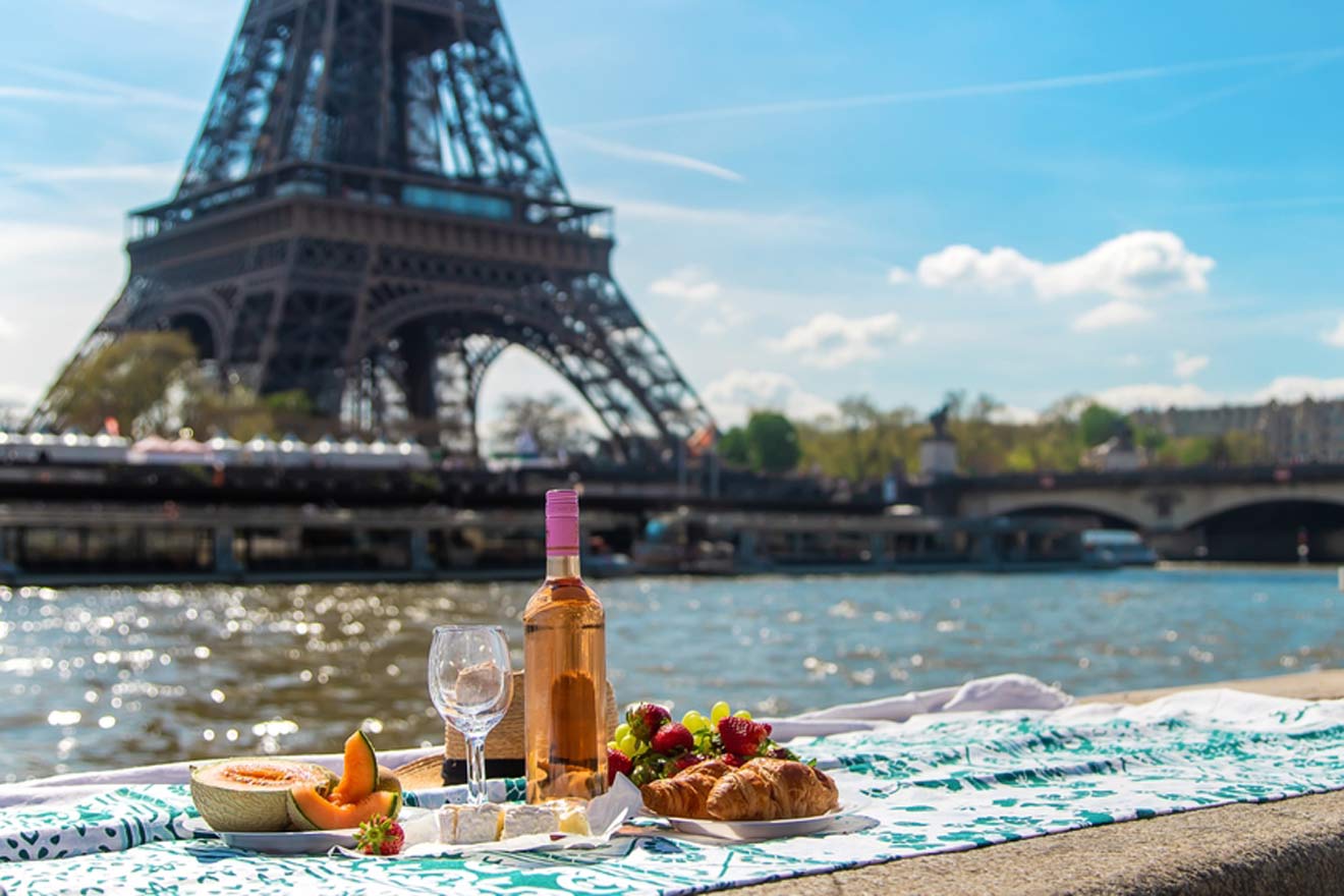 A picnic setup with wine, cheese, fruits, and a croissant on a riverside near the Eiffel Tower on a sunny day.