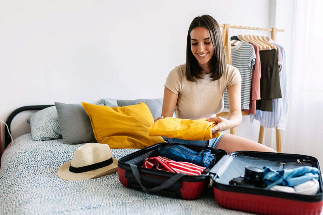 A woman sits on a bed packing a suitcase with clothes. A hat and more clothes are nearby. A clothes rack with hanging garments is visible in the background.