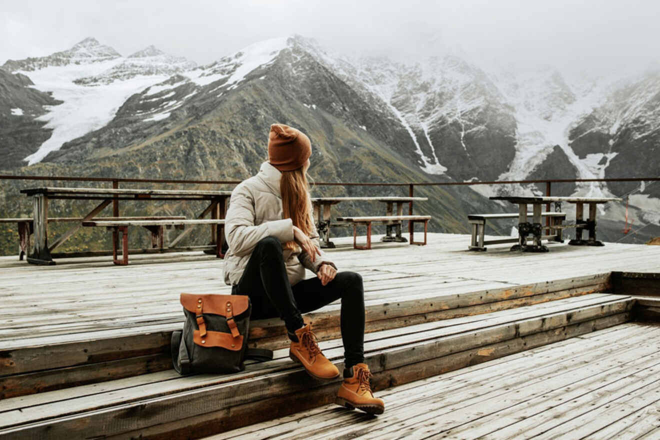 A person in a beanie and jacket sits on wooden steps with a backpack, surrounded by snowy mountains and overcast skies.