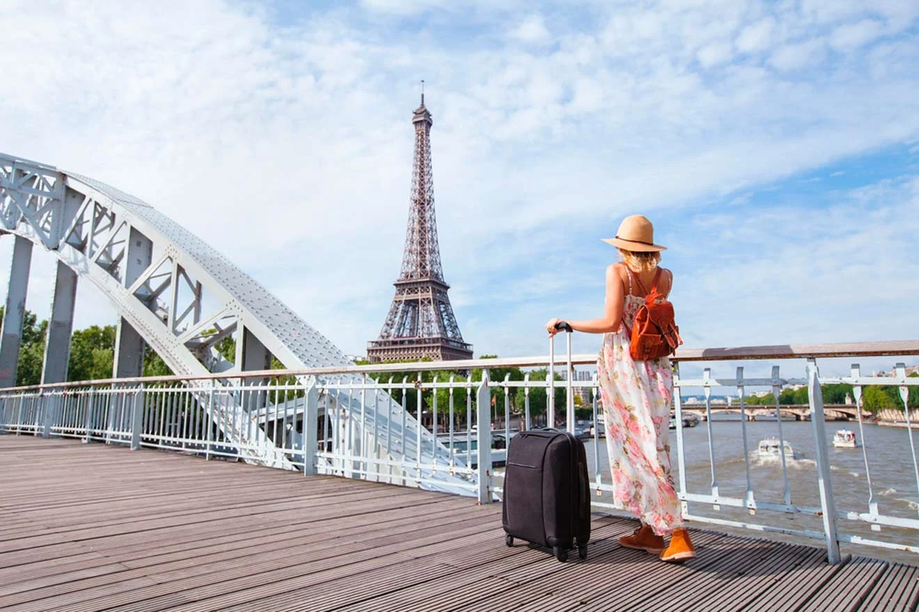 A woman in a summer dress and hat stands on a bridge with a suitcase, overlooking the Eiffel Tower in Paris on a sunny day.