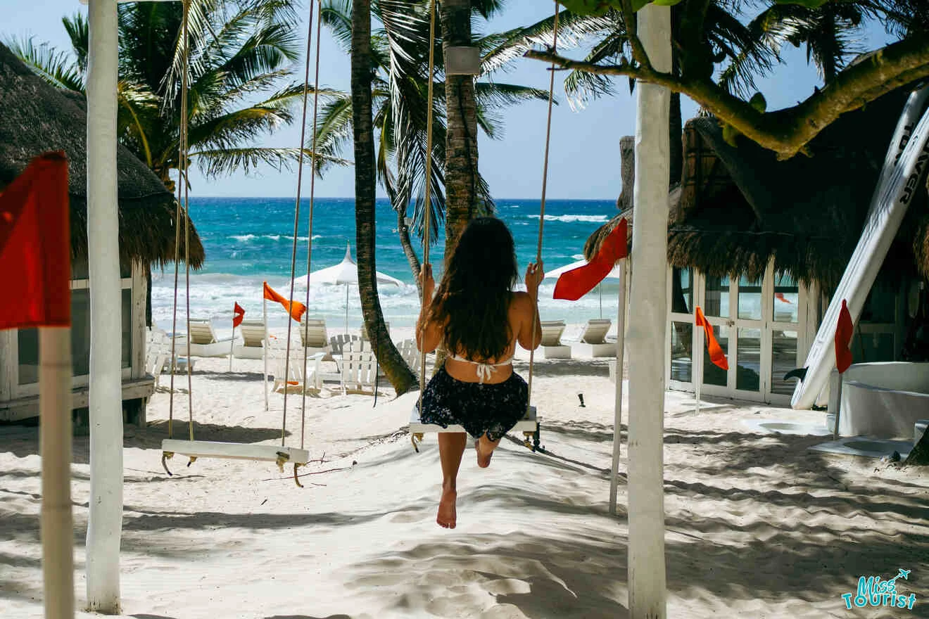 the author of the post with long hair sits on a swing facing the ocean at a beach resort surrounded by palm trees and huts, with red flags marking the area.