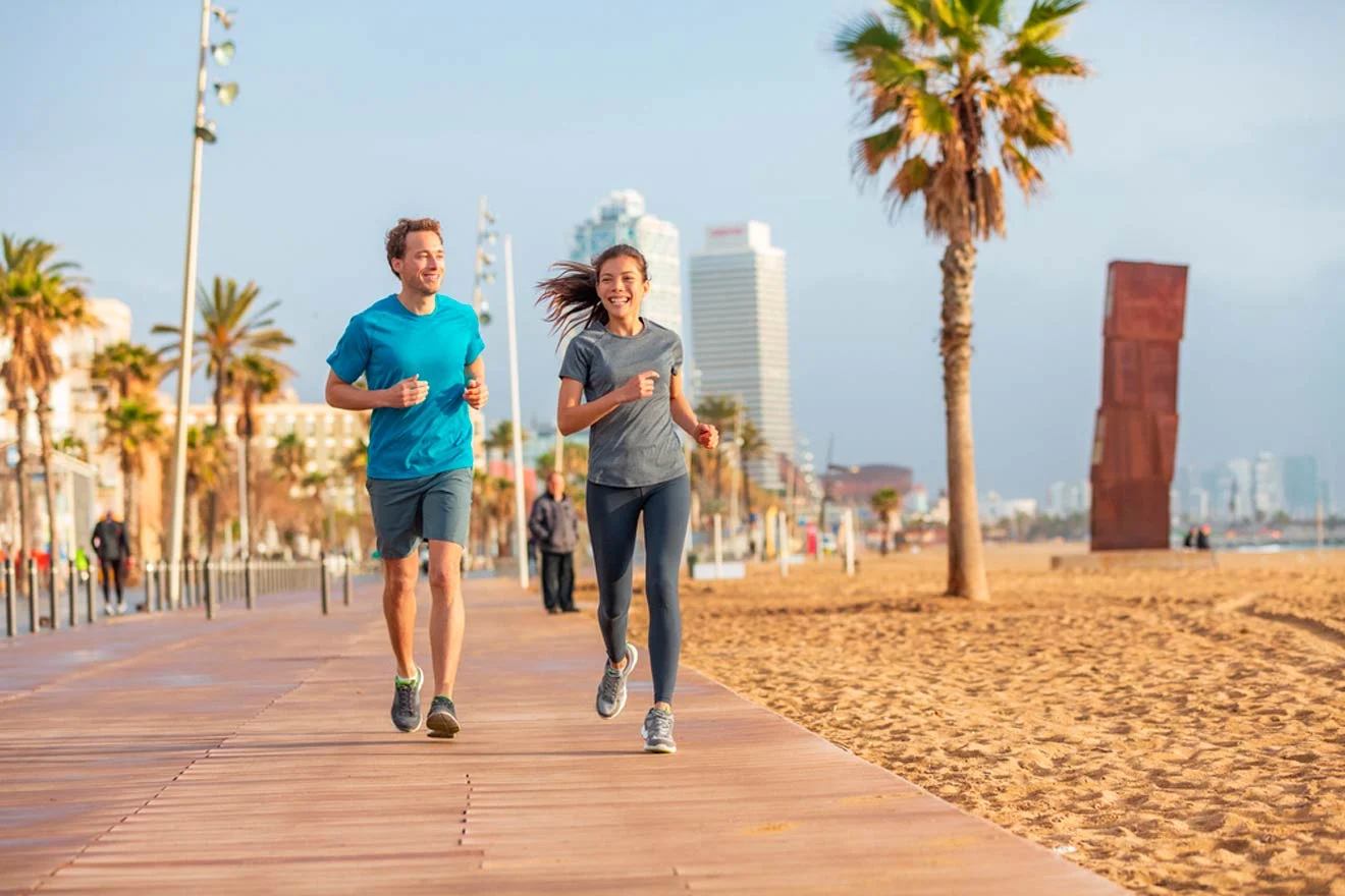 Two people are jogging on a wooden boardwalk by the beach, with palm trees and tall buildings visible in the background under a clear sky.