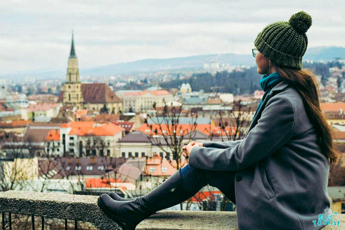 A person in a green hat and grey coat is sitting on a ledge overlooking a cityscape with a prominent church steeple and red rooftops.